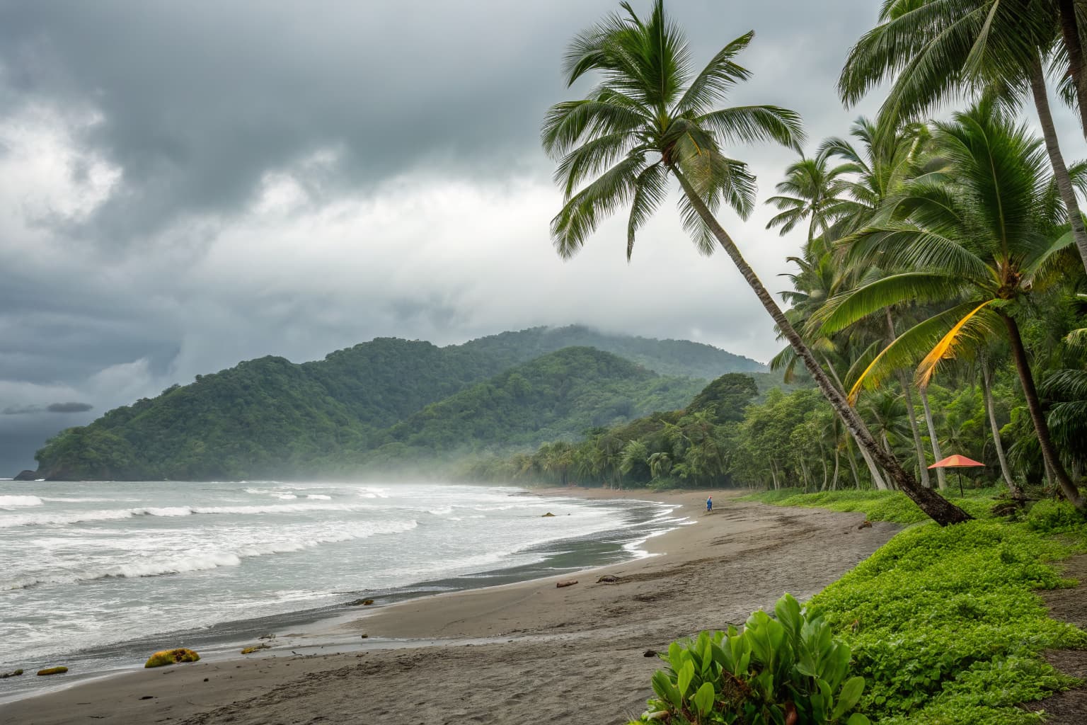 Jaco Beach Costa Rica during rainy season — lush green jungle hills draped in mist, palm trees along the Pacific shoreline under dramatic storm clouds