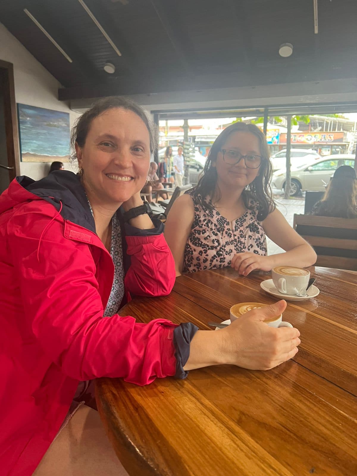 Two women sharing coffee at an open-air Jaco cafe during a rainy day, one wearing a red rain jacket